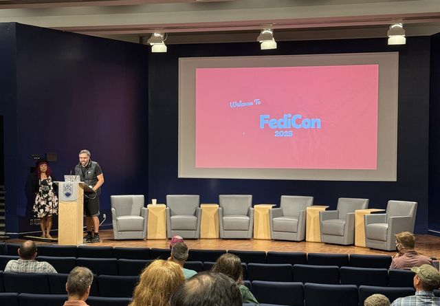 Photo of the stage at FediCon 2025. The organizer is giving a speech while an image saying 'Welcome to FediCon 2025' is projected