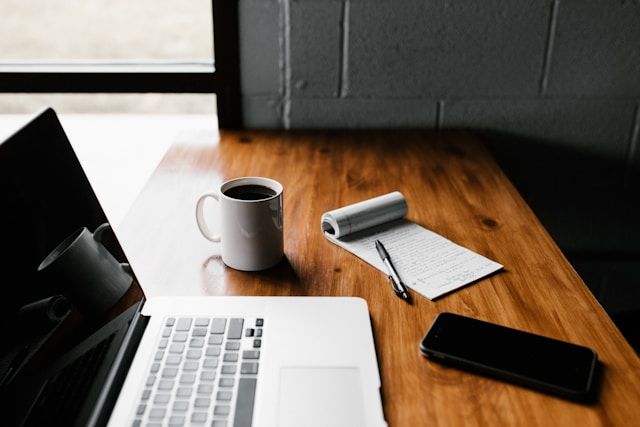 Picture of a desk. On top of it, there's a Macbook, a coffee mug, a phone and a notebook with a pen on top.
